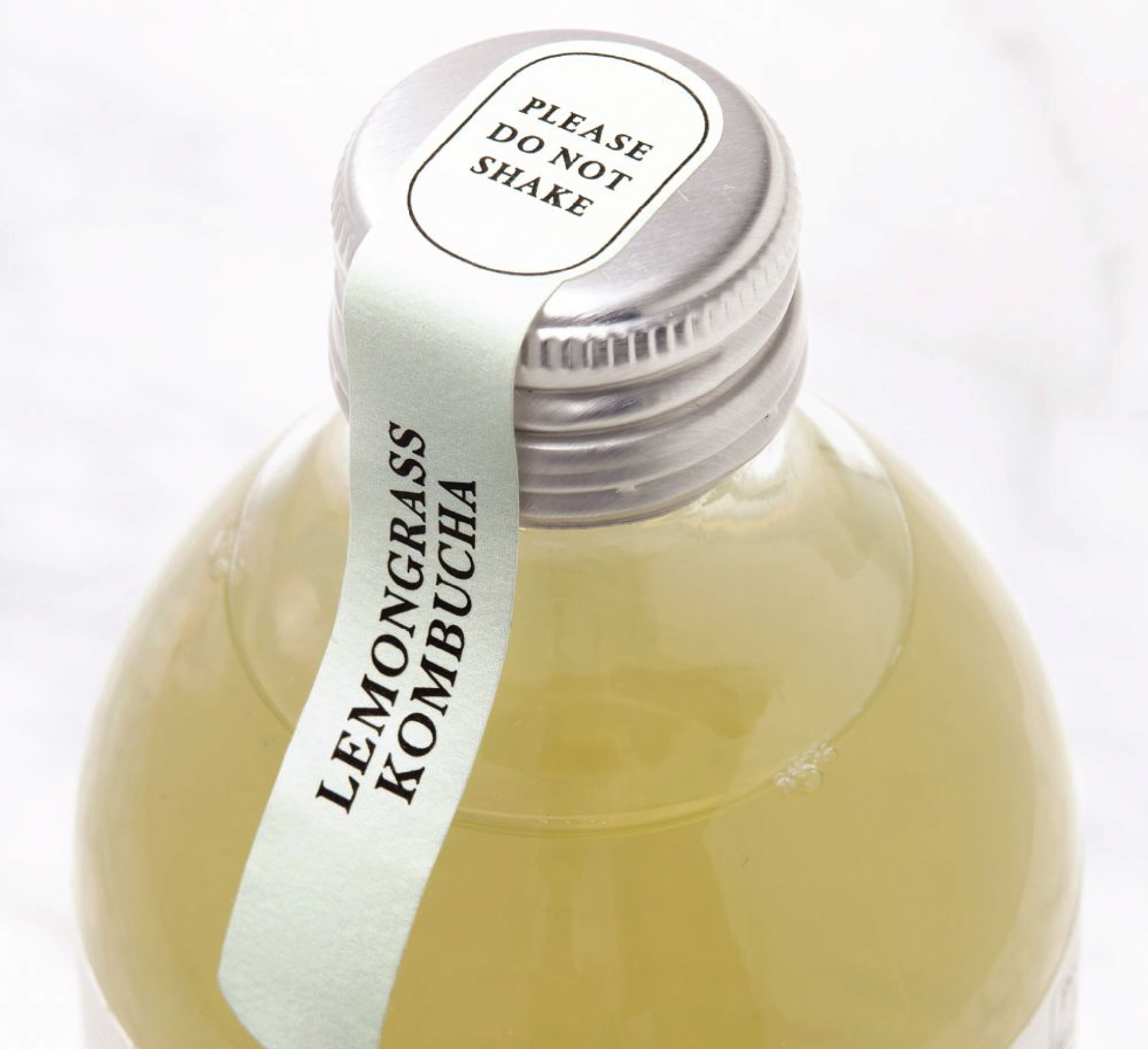 Pale green neck seal label extending over a silver bottle cap, printed with Lemongrass Kombucha and Please Do Not Shake.