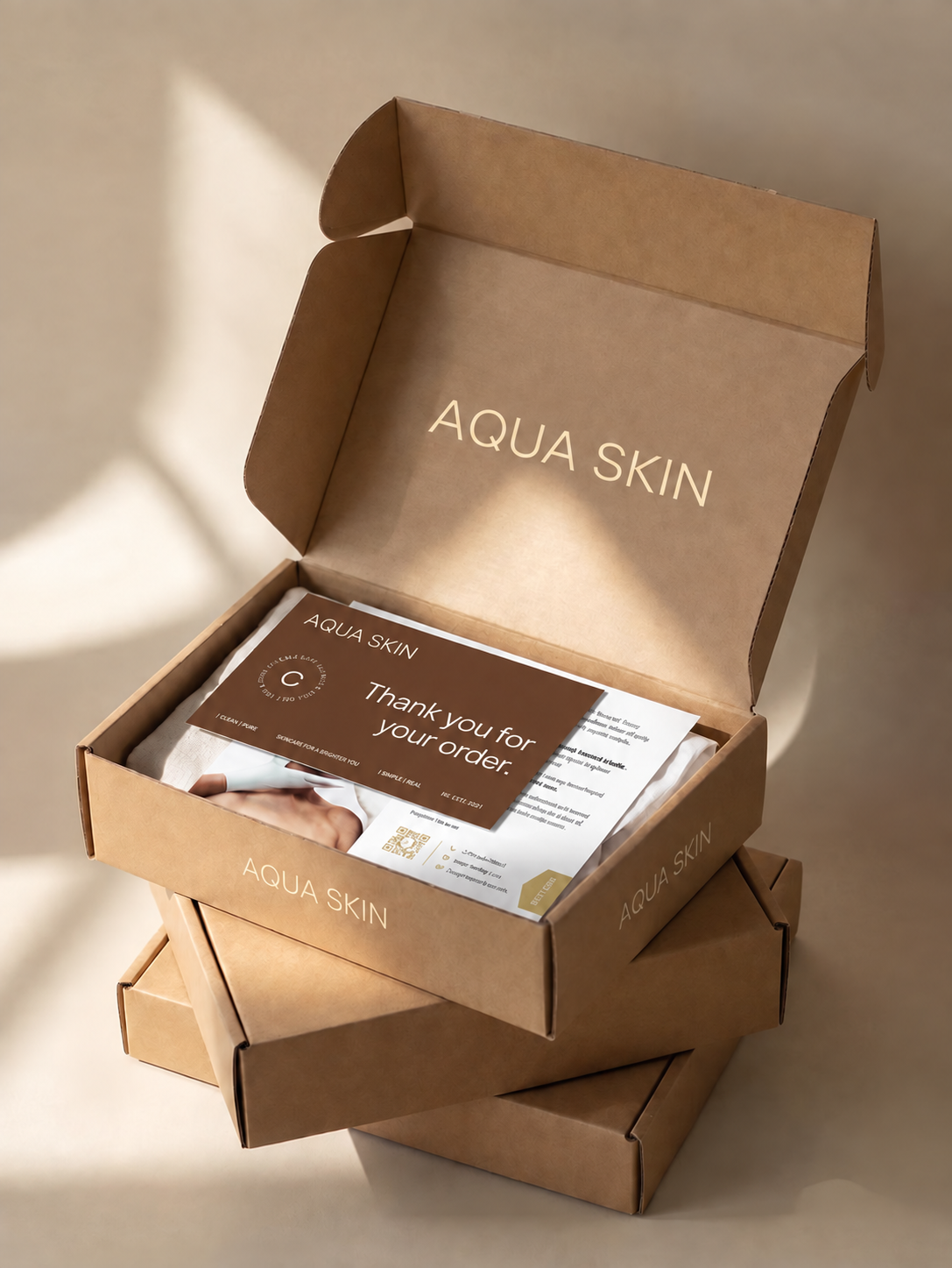 Open brown kraft mailer box with AQUA SKIN branding, a thank-you card and printed leaflet, stacked above closed boxes.