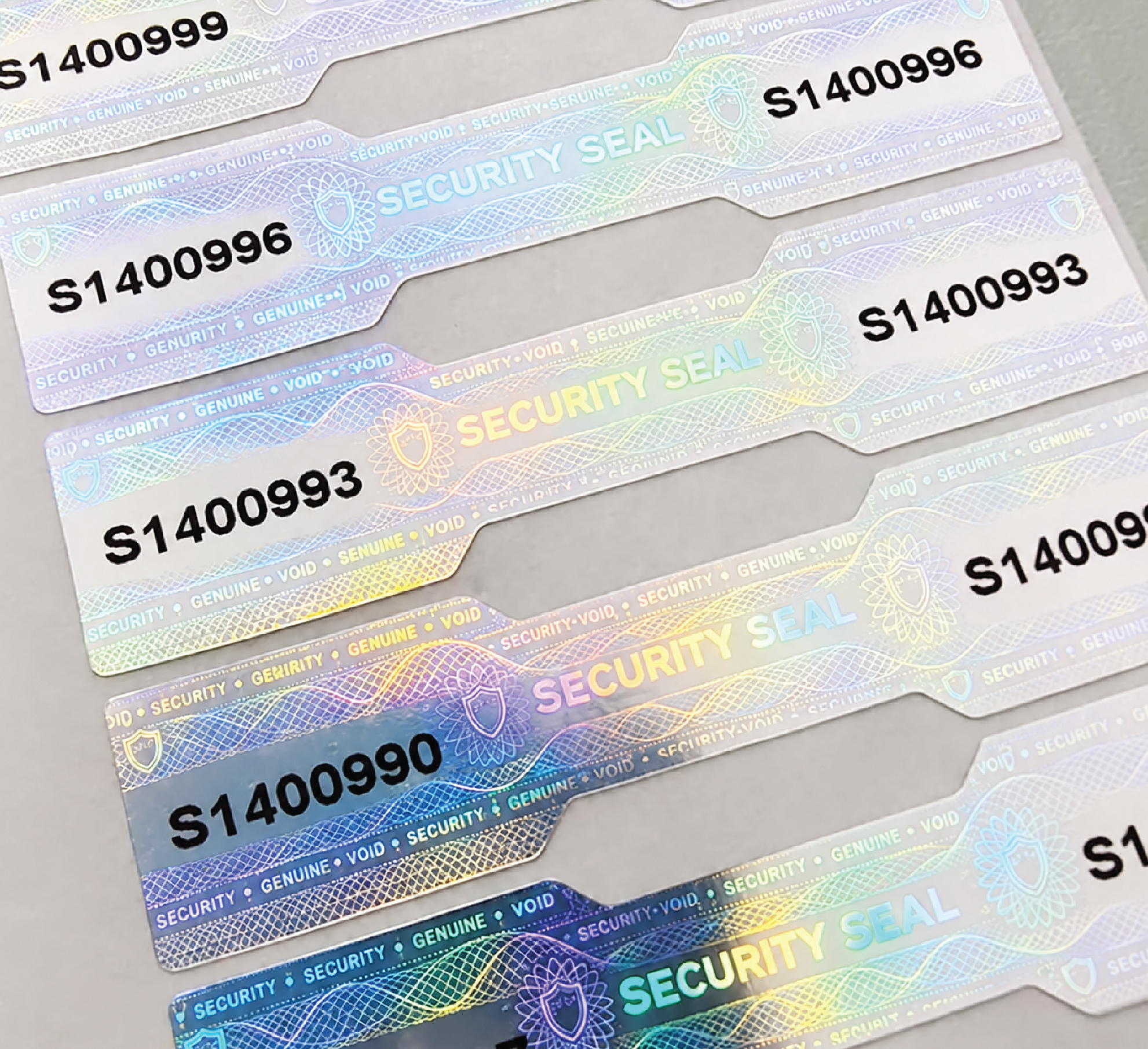 Silver holographic security seal labels with narrow centre sections, shield patterns and black serial numbers at both ends.