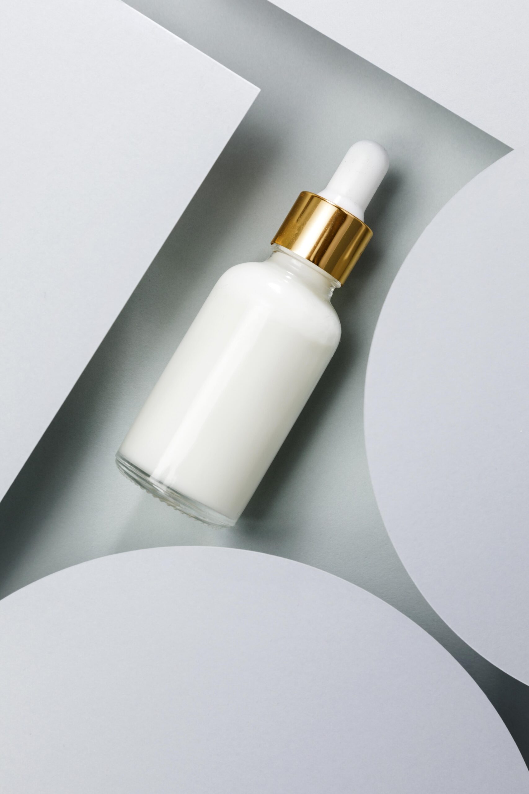 White cosmetic dropper bottle with a gold collar and white rubber bulb on a grey and white geometric background.